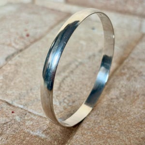May include: A shiny, silver-colored bangle bracelet. The bracelet has a smooth, rounded design and reflects light, creating highlights and shadows. The jewelry is displayed on a textured, light brown surface.
