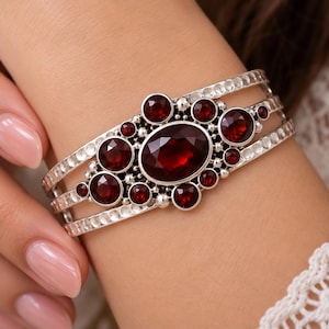 May include: A silver cuff bracelet featuring a central large oval faceted deep red garnet stone surrounded by smaller round faceted garnet stones. The metal band has a hammered texture and consists of three parallel silver bars connected together.