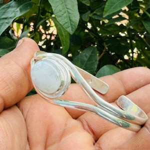Puede incluir: Un brazalete de plata con un cabujón de piedra lunar blanco translúcido de forma ovalada y lisa, engastado en un bisel de metal pulido. El brazalete tiene un diseño abierto con dos bandas de plata paralelas que se curvan alrededor de la gema.
