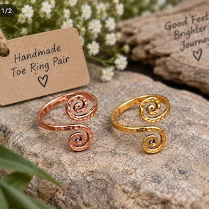 May include: A pair of open-ended toe rings featuring a hammered texture and spiral ends. One ring is copper-toned and the other is gold-toned. A tan tag is present with the text Handmade Toe Ring Pair and Good Feet Brighter Journeys.
