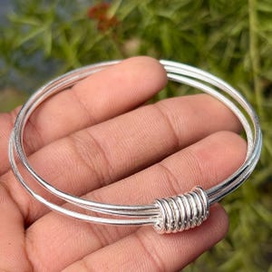 May include: A silver bangle bracelet with a coiled design. The bracelet is made of three thin, round bands of silver, held together by a coiled section. The bracelet is shown against a blurred green background.