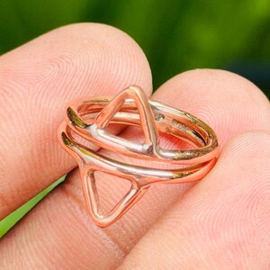May include: A rose gold-toned ring featuring two stacked bands, each topped with a prominent, open triangular geometric shape. The metal has a polished, reflective finish, and the design creates a layered appearance with the two triangles aligned.