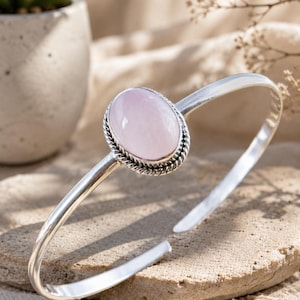 Pode incluir: Uma pulseira de prata com uma pedra preciosa de quartzo rosa claro, lisa e de formato oval, no centro. A pedra está engastada numa borda de prata com um design de corda torcida, e a banda é de metal prateado polido e aberto.