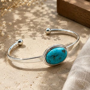 May include: A silver cuff bracelet featuring a polished metal band with a rounded finish and a single, large, oval-shaped turquoise stone set in the centre. The bracelet ends in two small, smooth silver spheres at the open gap of the cuff.