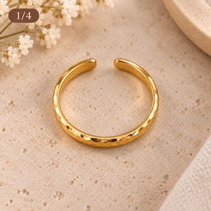 May include: A gold-toned open ring featuring a textured, hammered surface with a polished finish. The band is circular with a small gap at the top, showcasing a reflective metallic sheen and a consistent, repeating indented pattern along the exterior.