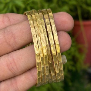Op de afbeelding: Een stapel van zes goudkleurige armbanden met een getextureerde, gehamerde afwerking. De armbanden zijn op elkaar gestapeld en tonen hun individuele ontwerpen en algehele esthetiek. De sieraden worden gepresenteerd tegen een vage achtergrond.