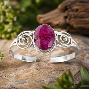 Può includere: Un bracciale a fascia in argento con una grande pietra preziosa sfaccettata di colore rosa intenso, di forma ovale, al centro. La fascia metallica è decorata con volute di filo simmetriche su entrambi i lati, creando un design aperto.