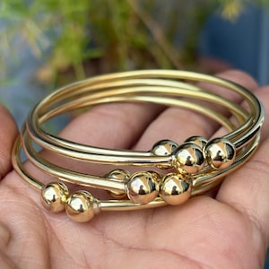 May include: A set of four gold-colored bangles. Each bangle has a smooth, rounded design with small, spherical accents. The bangles are stacked together, creating a layered effect. The jewelry is displayed against a blurred background of green foliage.