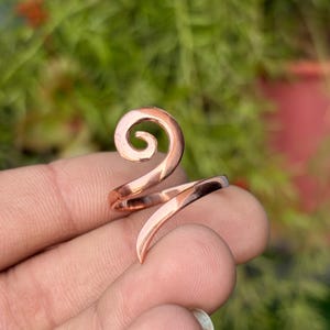 May include: A copper-coloured ring with a spiral design. The ring wraps around the finger with a curved, open-ended design. The ring is shown against a blurred green background.