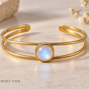 May include: A gold-toned cuff bracelet featuring a double-band design and a central circular setting. The setting holds a smooth, round moonstone cabochon that displays a soft blue iridescent glow. The text 1. FRONT VIEW is visible.