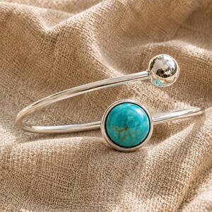 Op de afbeelding: Een zilverkleurige open armband met een gepolijst bolvormig uiteinde en een ronde zetting met een heldere turquoise steen met bruine aders. De metalen band is glad en reflecterend, wat zorgt voor een strak en modern uiterlijk.