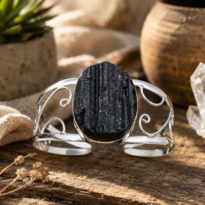 Op de afbeelding: Een zilverkleurige armband met een grote, ovale, ruwe zwarte toermalijnsteen in het midden. De metalen band is aan beide zijden voorzien van opengewerkte krulpatronen, die een decoratief frame vormen voor het donkere, getextureerde mineraal.