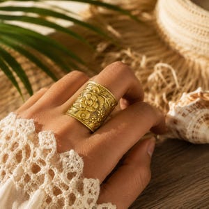 May include: A wide gold-toned metal ring featuring an embossed floral pattern with intricate vine and leaf details. The band has a textured finish and a straight edge, showcasing a repeating botanical design that wraps around the entire circumference.