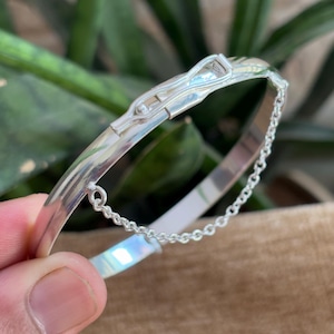 Peut inclure: Un bracelet jonc argenté avec un fermoir à charnière et une chaîne de sécurité. Le bracelet est incurvé et poli, reflétant la lumière. Le fermoir a un design décoratif. La chaîne est faite de petits cercles liés.