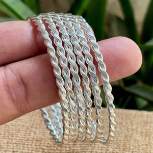 Può includere: Una collezione di braccialetti color argento con un design a corda intrecciata. I braccialetti sono impilati, mostrando il loro intricato motivo e la superficie riflettente. I gioielli sono esposti su uno sfondo neutro.