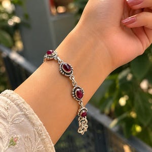 May include: A silver chain bracelet featuring four oval-shaped, deep red cabochon gemstones set in ornate metal bezels. The bracelet includes a decorative metal knot charm at the end and connects with a series of small, polished silver links.