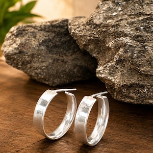 Peut inclure: Une paire de boucles d'oreilles créoles argentées. Les boucles d'oreilles ont un design plat et large avec une finition polie. Elles sont présentées sur une surface en bois avec un arrière-plan en pierre naturelle.