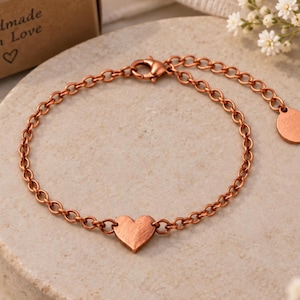 May include: A copper-toned metal chain bracelet featuring a central heart-shaped charm and a lobster claw clasp. The bracelet includes an adjustable extension chain ending in a small round metal tag. The surface of the heart charm has a brushed texture.