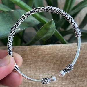 Op de afbeelding: Een zilverkleurige open manchetarmband. De armband heeft een gedetailleerd ontwerp met een reeks met elkaar verbonden S-vormen en geweven texturen. De uiteinden van de armband hebben kleine zilveren bollen en geweven details.