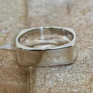 Può includere: Primo piano di un anello d'argento con una fascia quadrata. L'anello ha una superficie liscia e lucida e un design semplice e moderno. L'anello è probabilmente realizzato in argento sterling o metallo simile.