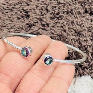 May include: A silver cuff bracelet with two iridescent gemstones. The gemstones show green, purple, and blue colours. The bracelet has an open design, with the gemstones set at each end. The bracelet is made of silver.