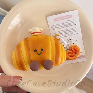 Puede incluir: Un accesorio amarillo en forma de croissant con gorro de chef y cara sonriente. Pies circulares marrones están unidos a la parte inferior. También se ve un pequeño pastel en forma de corazón y una etiqueta con texto.