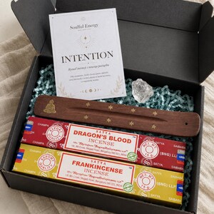 Pode incluir: Uma caixa de presente preta contendo duas caixas retangulares de incenso Satya nos aromas Dragon's Blood e Frankincense, um queimador de incenso de madeira com motivos de estrelas douradas e Buda, um cristal transparente e um cartão branco com o texto Soulful Energy INTENTION.