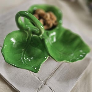 Könnte beinhalten: Eine hellgrüne, blattförmige Keramikservierschale mit Griff. Die Schale hat drei Abschnitte, von denen einer Nüsse enthält. Die Schale steht auf einer weißen Leinen-Serviette. Die glänzende Oberfläche der Schale reflektiert das Licht.