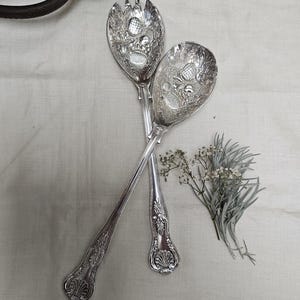 Könnte beinhalten: Ein Set aus drei Keramikbechern mit gesprenkelter, cremefarbener Glasur. Jeder Becher zeigt ein handgemaltes Design: ein blau-grünes Blatt, eine gelbe Blume und eine rote Beere. Die Becher haben eine einfache, abgerundete Form.