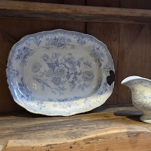 Könnte beinhalten: Eine ovale Keramikplatte mit blau-weißem Blumenmuster, gewelltem Rand und einer dunklen Absplitterung an der rechten Seite. Daneben steht eine passende Keramik-Sauciere mit blau-weißem Blumenmuster auf cremefarbenem Untergrund.
