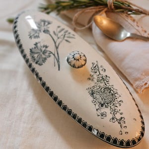 Puede incluir: Una fuente ovalada de cerámica blanquecina con tapa, con diseños florales y geométricos negros. La tapa tiene un pequeño pomo redondo. Una ramita de romero y una cuchara en el fondo sugieren un ambiente de comedor.