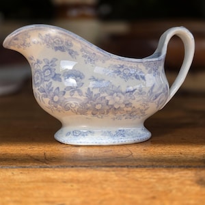 May include: A ceramic gravy boat featuring a cream-coloured base with a blue floral pattern. The vessel has a curved pouring spout, a single handle on the side, and a pedestal base. The intricate blue botanical design covers the exterior surface.