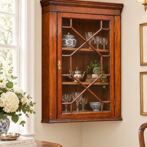 May include: A wall-mounted corner cabinet made of polished brown wood with a single glass door featuring a decorative lattice overlay. The interior contains two wooden shelves displaying various glass stemware, a silver teapot, and ceramic bowls.