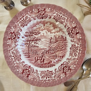 Puede incluir: Un plato redondo de estilo antiguo con un diseño de transferencia rojo y blanco. El plato presenta una escena de paisaje detallada con un castillo, árboles y caballos. El borde está decorado con patrones florales y de desplazamiento. Se ven cubiertos y saleros y pimenteros de plata.