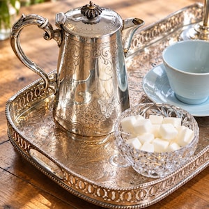 Könnte beinhalten: Eine versilberte Kaffeekanne mit verzierten Blumengravuren und einem dekorativen Knauf, platziert auf einem passenden rechteckigen Silbertablett mit aufwendigem Rand. Eine Kristallglasschale mit weißen Zuckerwürfeln steht auf dem Tablett.