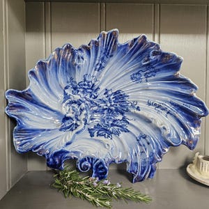 May include: A large, ornate, blue and white ceramic serving dish with a floral design. The dish has a ruffled edge with gold trim. A sprig of rosemary rests on the surface below the dish.