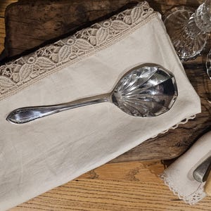 Puede incluir: Una cuchara de servir plateada con un cuenco facetado descansa sobre una servilleta de lino color crema con ribete de encaje. La cuchara está sobre una superficie de madera, con un candelabro y cubiertos en el fondo.