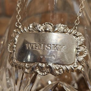 以下が含まれることがあります： クリスタルウイスキーデカンタのクローズアップ。シルバーのラベルには「WHISKY」と書かれており、装飾的なボーダーが付いています。ラベルはシルバーチェーンでデカンタに固定されています。デカンタはカットガラスのデザインです。