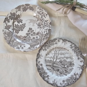 Puede incluir: Dos platos decorativos con una escena pastoral en blanco y negro. Los platos presentan un paisaje detallado con árboles, casas y un puente. Los platos están sobre un mantel crema, con una servilleta y cubiertos.