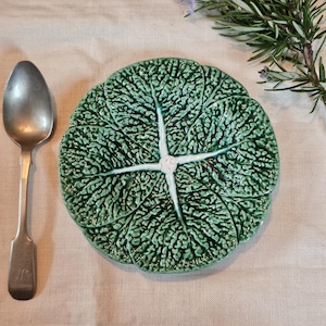 May include: A green ceramic plate shaped like a cabbage leaf, with a silver spoon on a neutral linen cloth. The plate has a textured surface and a white centre. A sprig of rosemary with purple flowers is in the top right corner.