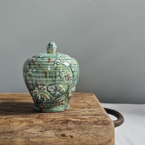Op de afbeelding: Een kleine, groene keramische pot met een deksel, versierd met een bloemenreliëfpatroon. De pot heeft een ronde vorm en staat op een houten oppervlak. Het kleurenpalet omvat groen, bruin en wit.