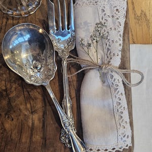 Peut inclure: Une cuillère et une fourchette argentées ornées de motifs floraux reposent sur un plateau en bois, à côté d'une serviette en lin pliée avec une bordure en dentelle. Une petite brindille de fleurs séchées est attachée à la serviette avec de la ficelle. Un couvercle en verre est en arrière-plan.