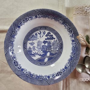 May include: A white and blue ceramic serving bowl with a classic Willow pattern. The bowl features a detailed blue landscape scene in the center, surrounded by a decorative border. The rim has a geometric design. A timeless piece for serving.