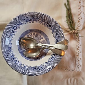 Puede incluir: Un cuenco de cerámica con estampado azul y blanco con dos cucharas de plata en su interior. El cuenco tiene un diseño detallado alrededor del borde y el centro. Una ramita de vegetación y un paño ribeteado de encaje están en el fondo.