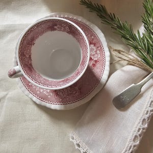 May include: A vintage tea cup and saucer set with a floral pattern in shades of pink and white. The cup and saucer are placed on a white linen surface, with a matching plate in the background. A white napkin with lace trim and a silver spoon are also present.