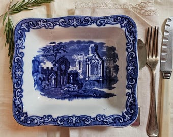 May include: A rectangular blue and white ceramic dish with a detailed landscape scene. The dish has a decorative border and is placed on a cream-colored surface with silverware and a sprig of rosemary.