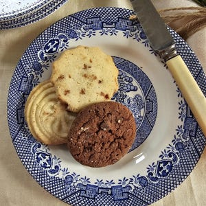 May include: A blue and white patterned plate holds three cookies: a light-colored, round cookie with nuts, a swirled cookie, and a chocolate chip cookie. A knife with a cream-colored handle rests on the plate. Another plate is partially visible.