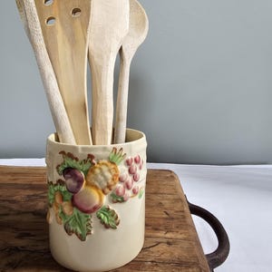Può includere: Un portautensili in ceramica color crema con un design a rilievo di frutta, contenente diversi cucchiai e spatole di legno. Il design della frutta comprende uva, prugne e pesche. Gli utensili in legno variano per forma e dimensione.