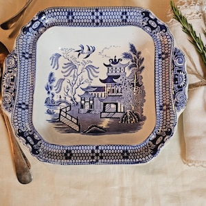 May include: Square blue and white ceramic serving dish with a detailed Willow pattern. The dish features a pagoda, bridge, and trees, with a decorative border. Silver serving utensils are on the side.