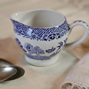 Puede incluir: Una cremera de cerámica blanca y azul con asa. La cremera presenta un diseño pastoral azul y blanco alrededor del cuerpo y el borde. La cremera está sobre una superficie blanca. Dos cucharas están a la izquierda de la cremera.
